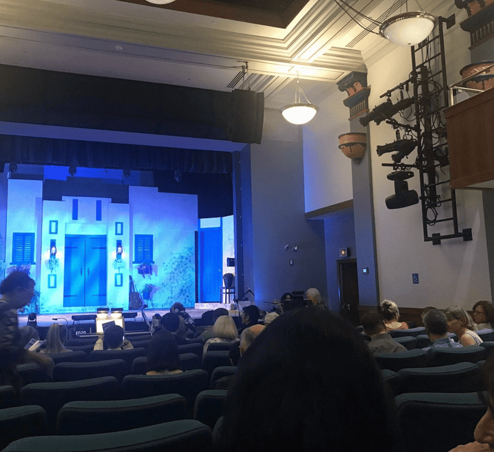 Elevated indoor theater scene with audience, spotlight, stage set, and technical equipment.