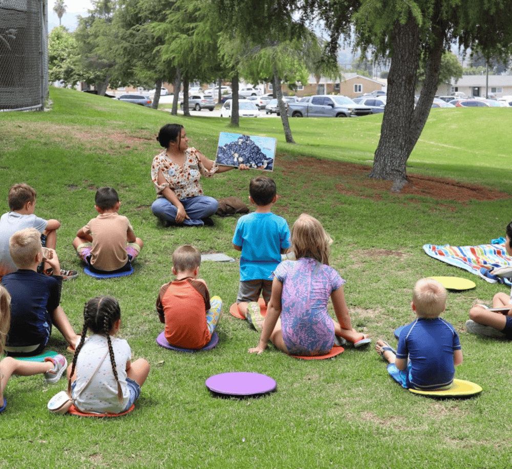Kids outdoor storytelling session at park with teacher, diverse children, educational activity, nature, community engagement, QuestForDirections.