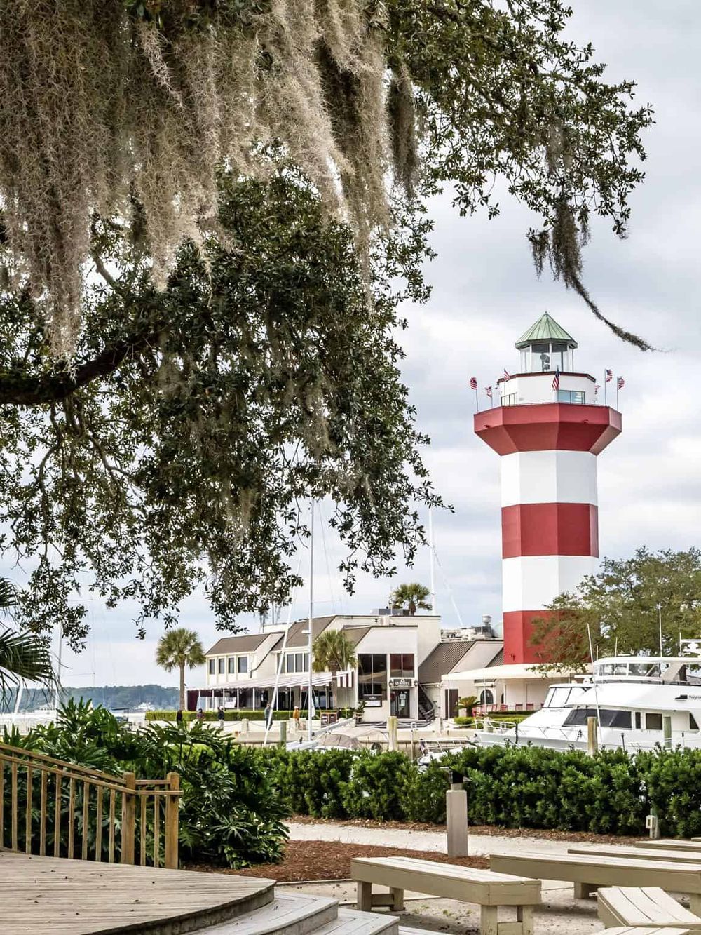 Historic lighthouse on waterfront with boats and lush greenery, a scenic coastal attraction.