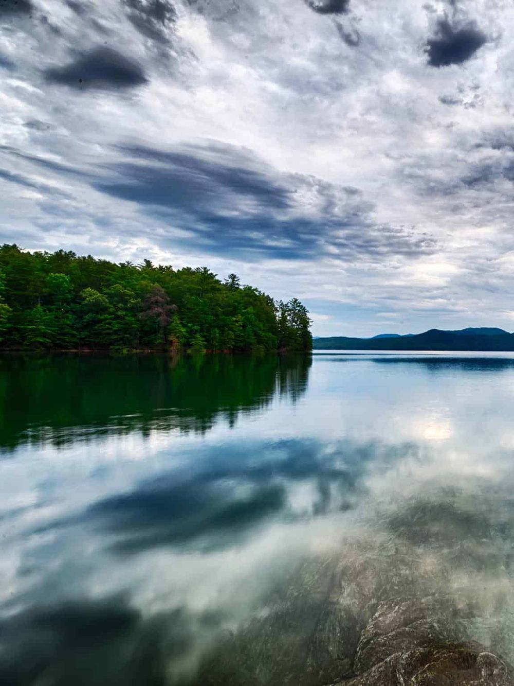 Serene lake with lush green shoreline and dramatic cloudy sky, perfect for travel and outdoor adventures.