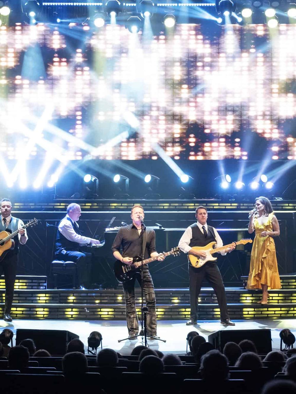 Bright live music performance with a full band on stage, colorful lights, and enthusiastic audience.