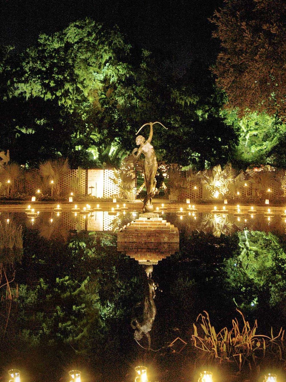 Serene nighttime garden with illuminated sculpture, lush trees, and reflective water feature for outdoor relaxation.