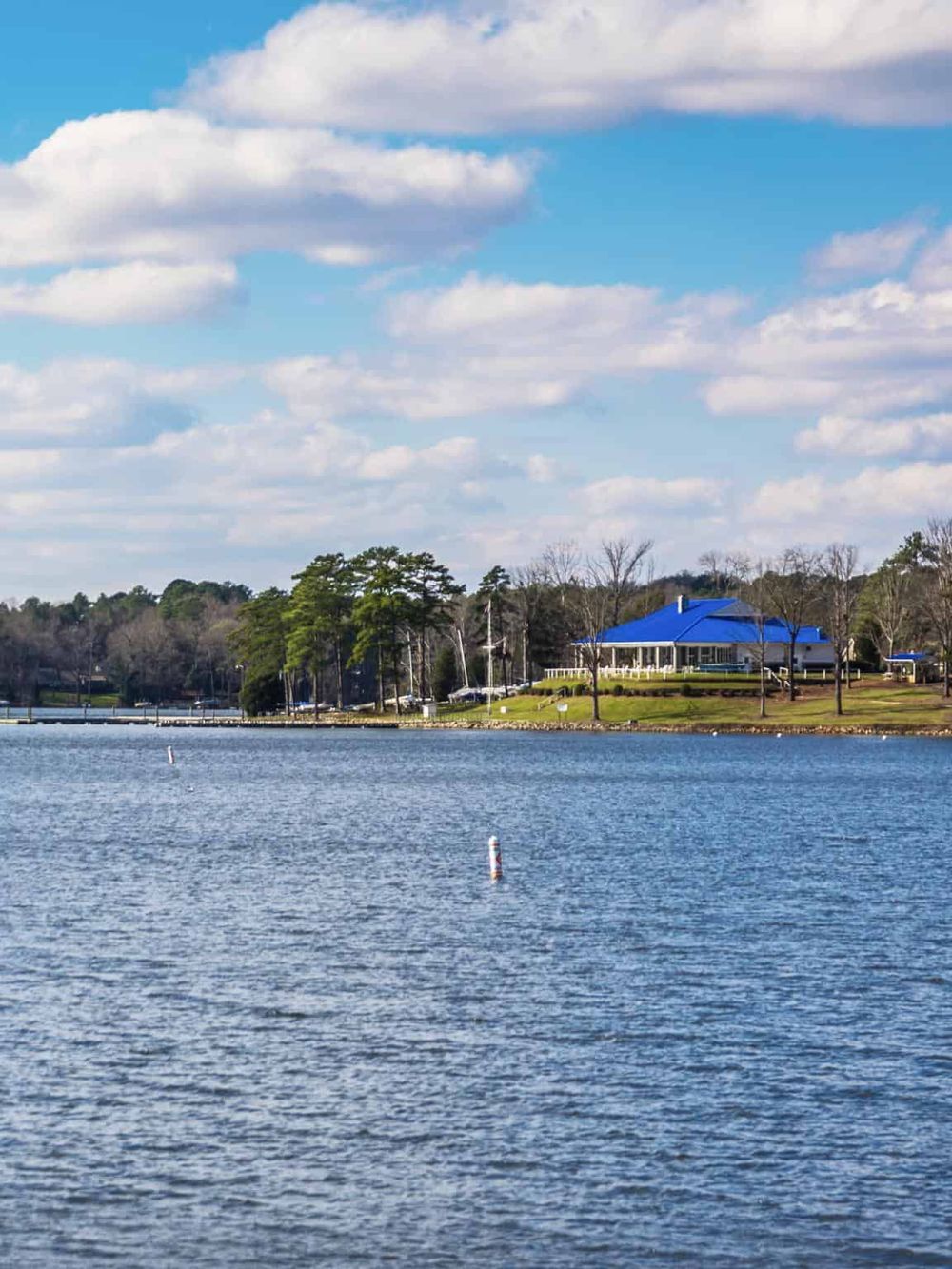 Serene lakeside scene with a building featuring a bright blue roof, surrounded by trees and calm water under a partly cloudy sky.