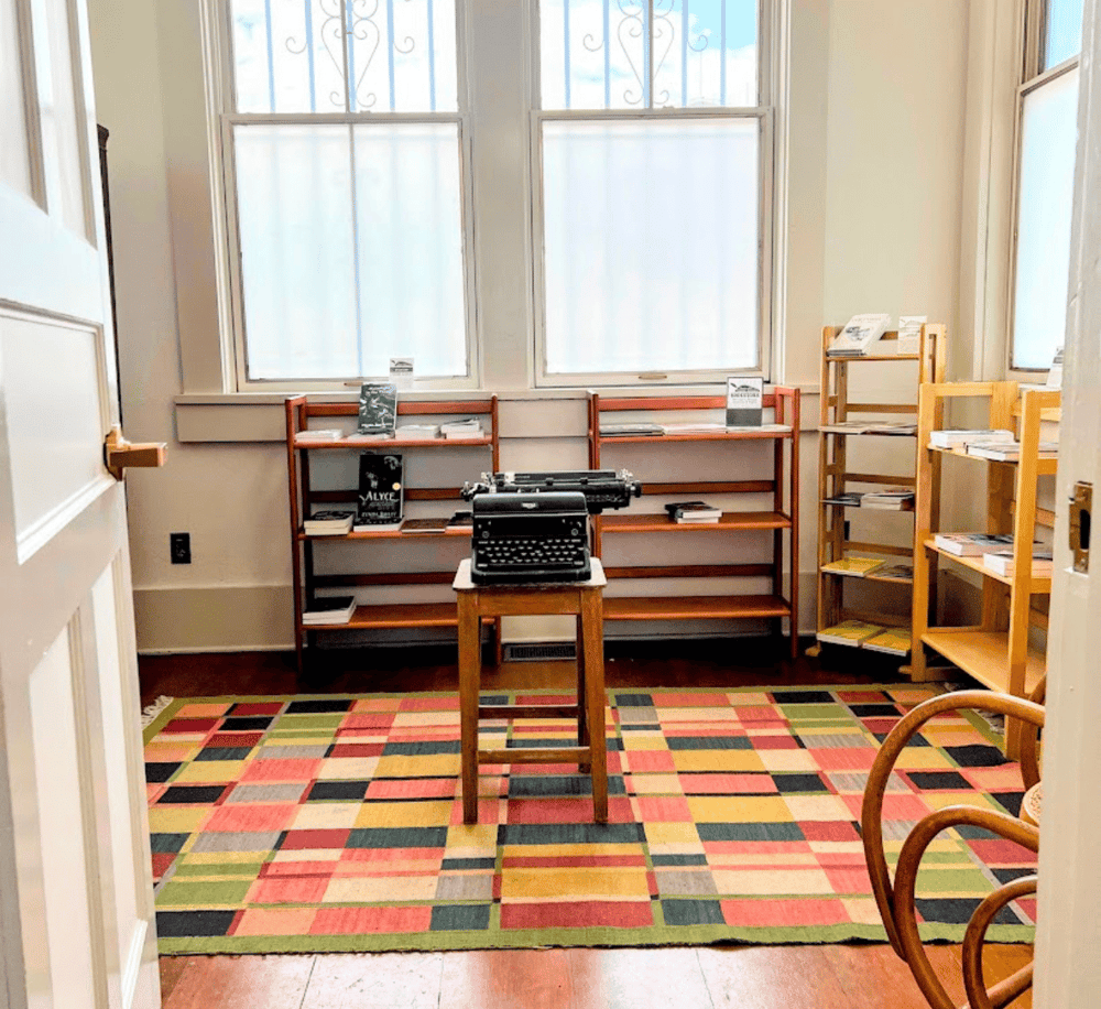 Typewriter on wooden table in cozy library corner with bookshelves, natural light, and colorful rug.