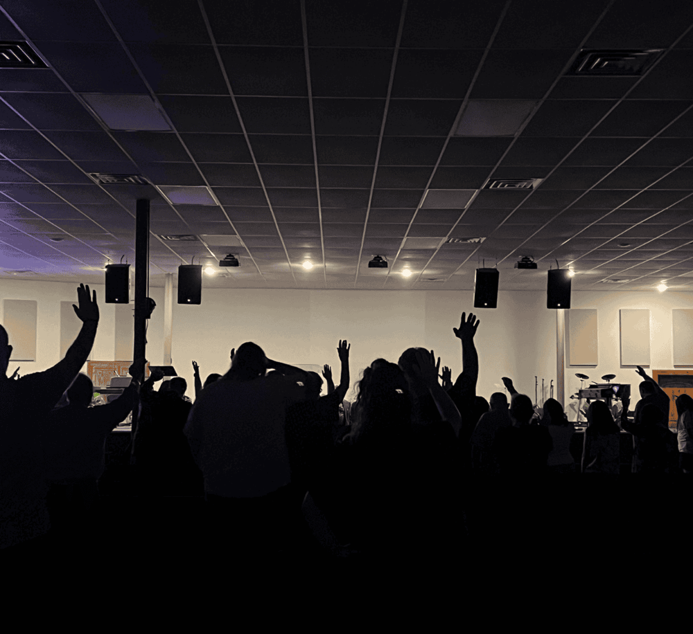 People raising hands in worship or concert audience with stage and speakers in background.