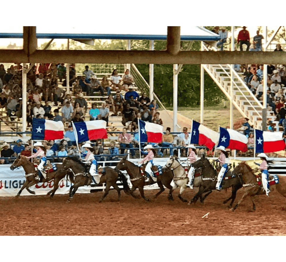 Colorful rodeo event featuring riders with Texas flags on horseback, lively crowd, and stadium stands.