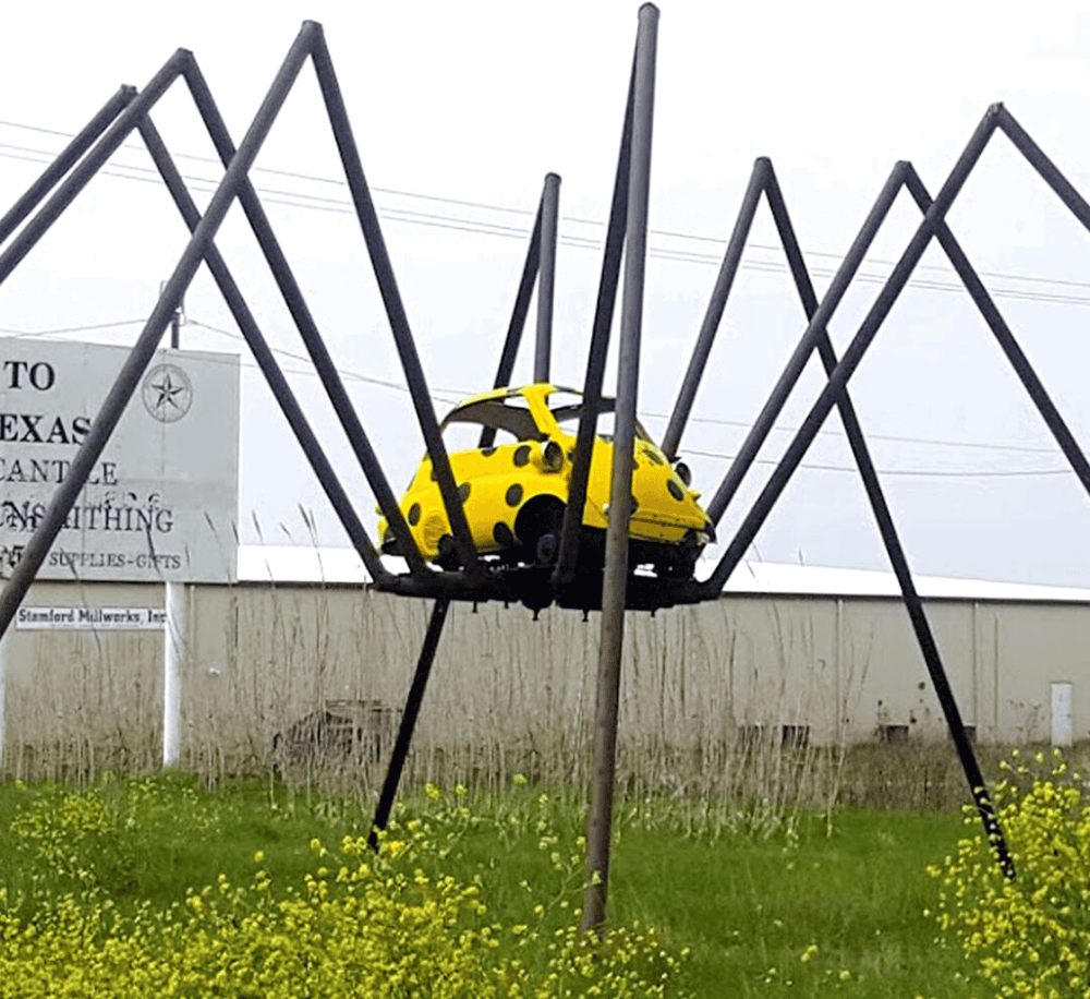 Tiny yellow car atop a decorative spider sculpture, symbolizing unique roadside attractions.