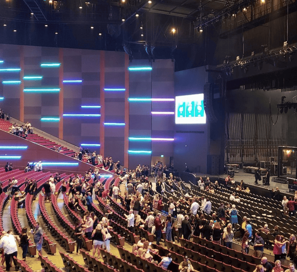 Bright indoor concert venue with a modern stage and vibrant LED lighting, featuring a seated audience waiting for the event to start.