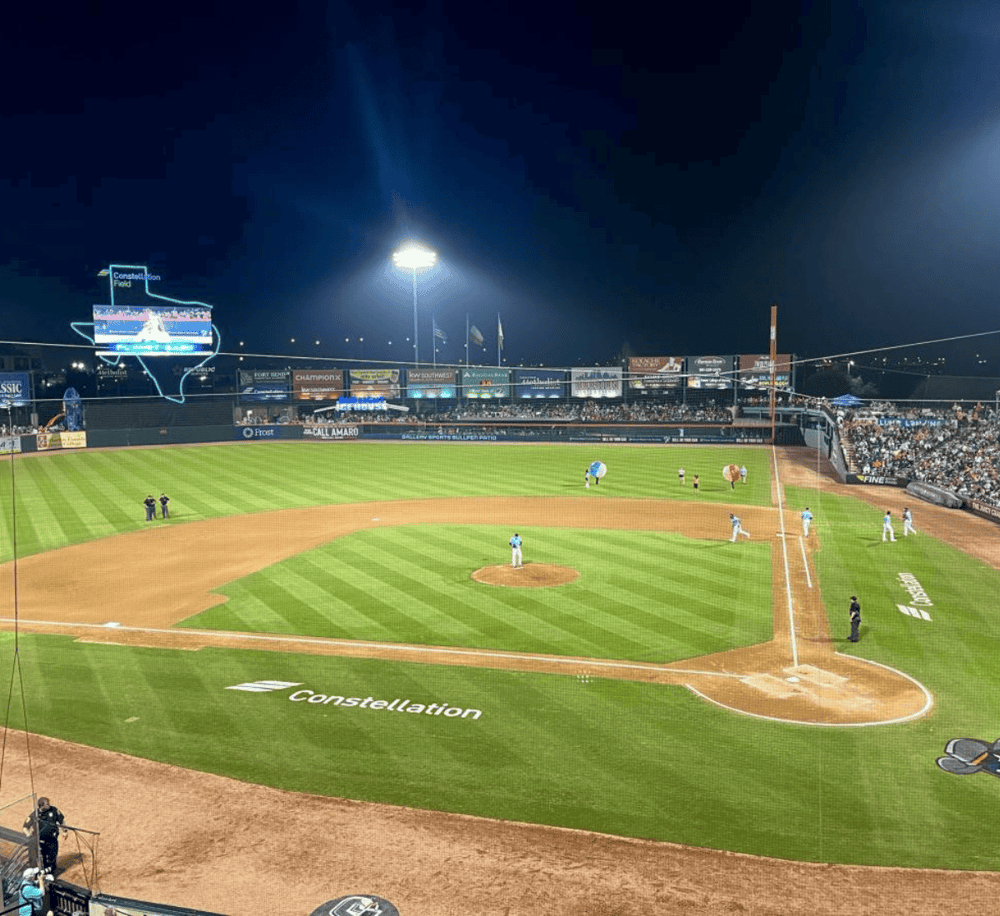 Brightly lit baseball stadium at night with players on the field and a large scoreboard in the background.