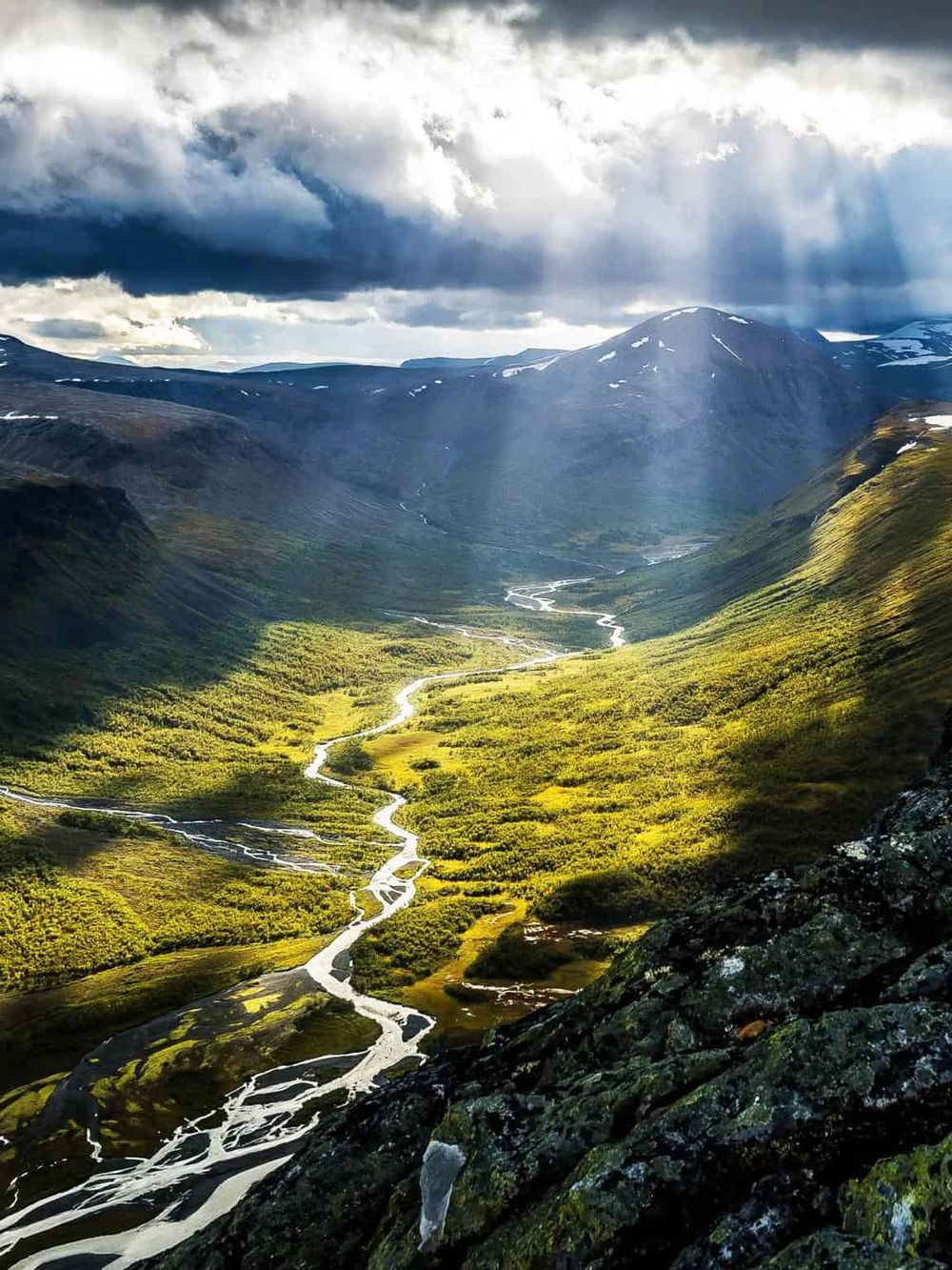 Breathtaking mountain valley with a winding river and lush greenery under dramatic cloud cover.