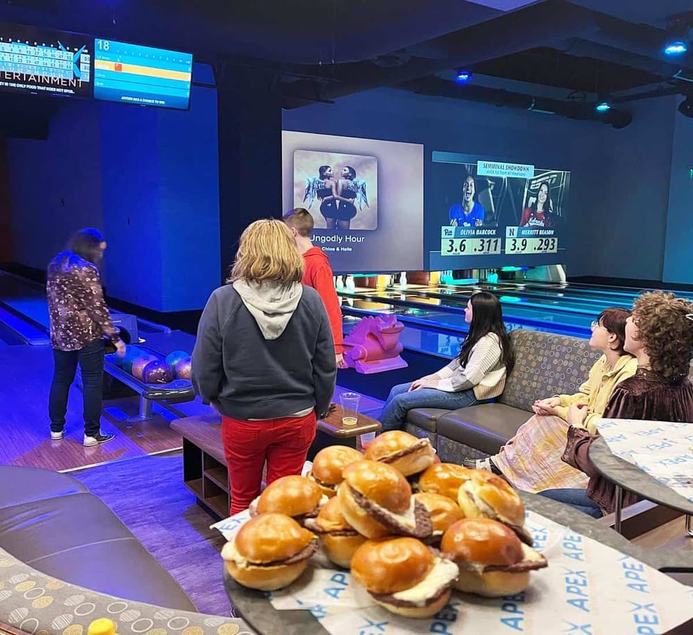 Astrip bowling alley with people watching a game on the big screen, food, and snacks.