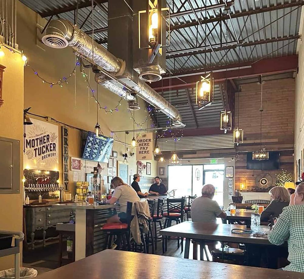 Bright brewery interior with patrons enjoying drinks, industrial decor, and festive string lights.