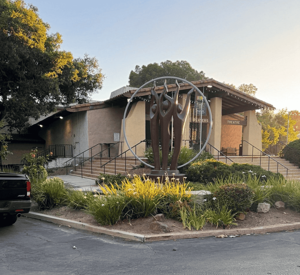 Artistic sculpture outside a community theater building, surrounded by landscaped greenery.