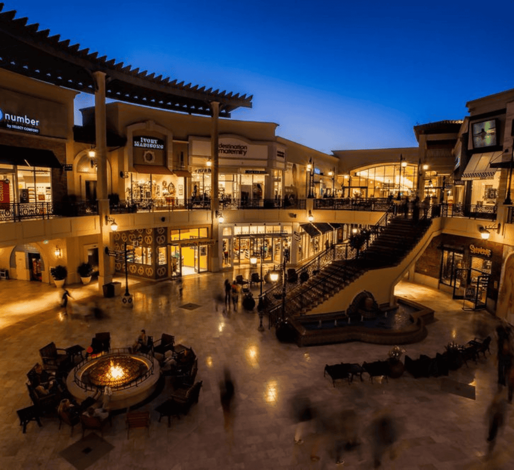 Bright indoor shopping mall with multi-level stores, warm lighting, and a central fire pit seating area.