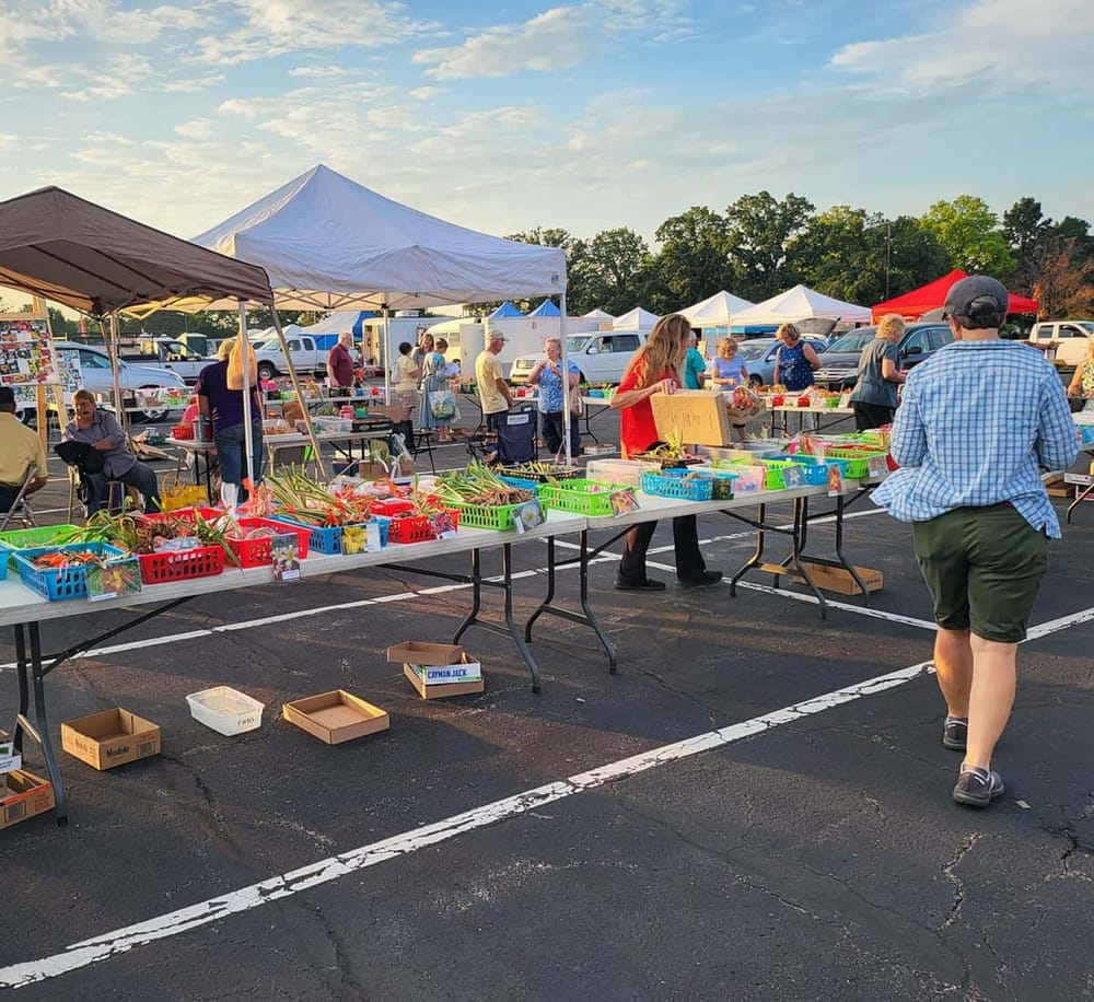 Colorful outdoor farmers market with vendors, shoppers, and tents in a parking lot.