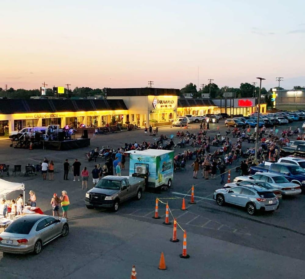 Outdoor shopping mall parking lot during evening with people gathering, store signage, and cars.