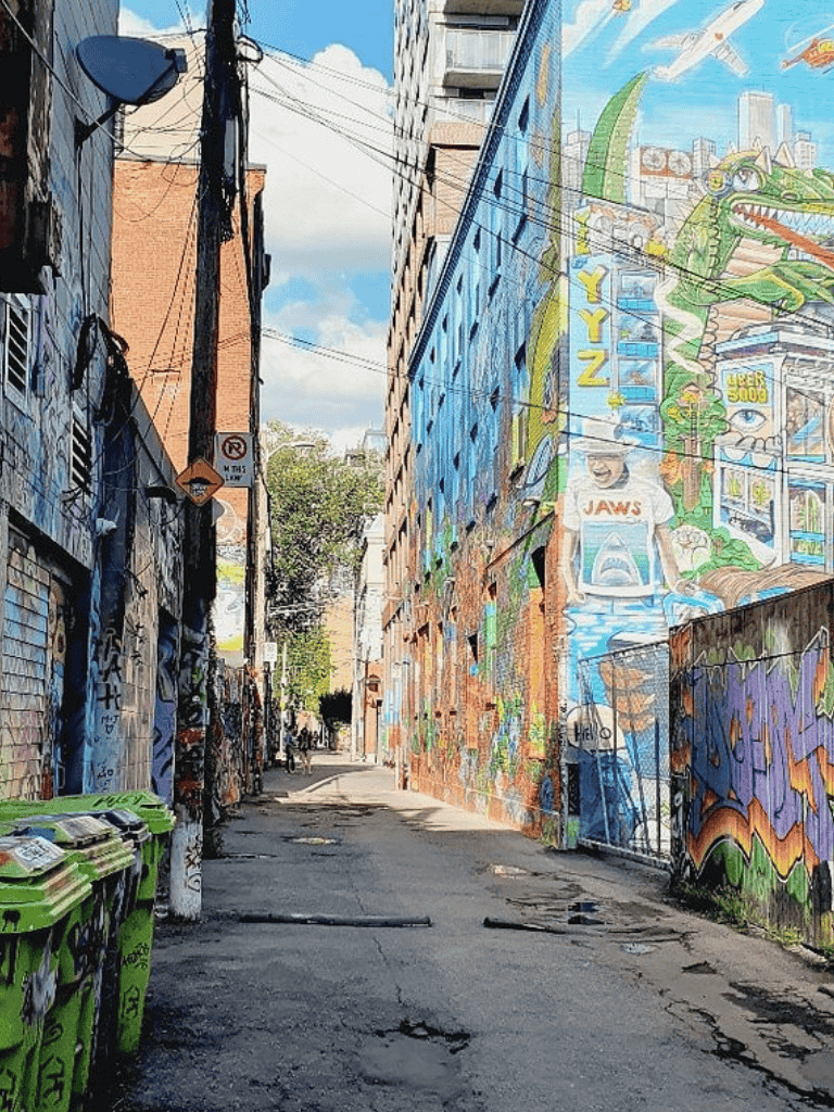 Colorful urban alleyway with vibrant mural art, graffiti, and cityscape in the background.