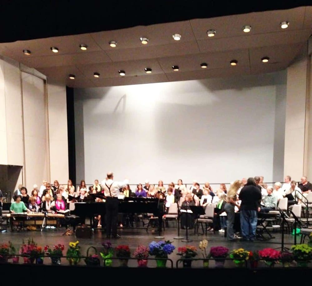 Vocal ensemble rehearsal on stage with conductor, colorful flowers in front, theater ambiance, QuestForDirections event.