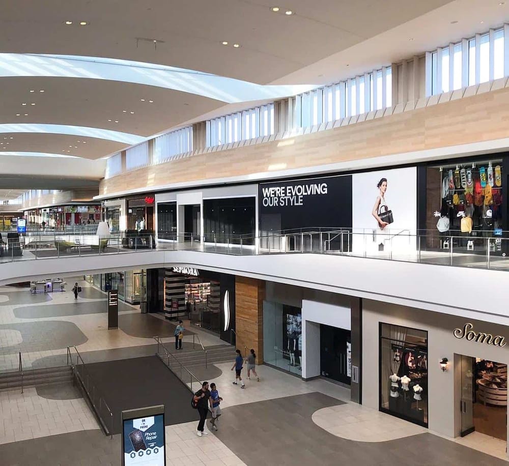 Bright indoor shopping mall with multiple store fronts and large digital advertising screens.