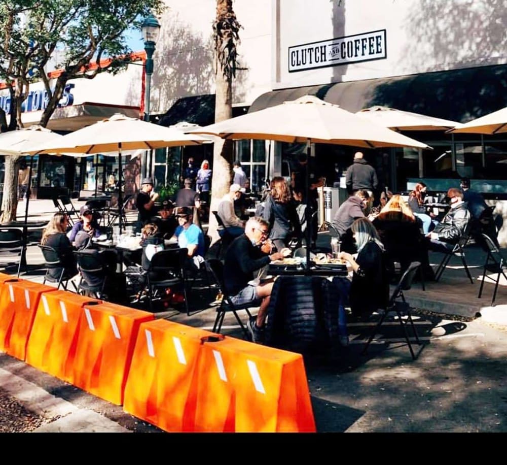 Outdoor coffee shop with people enjoying drinks under umbrellas, bustling city atmosphere.