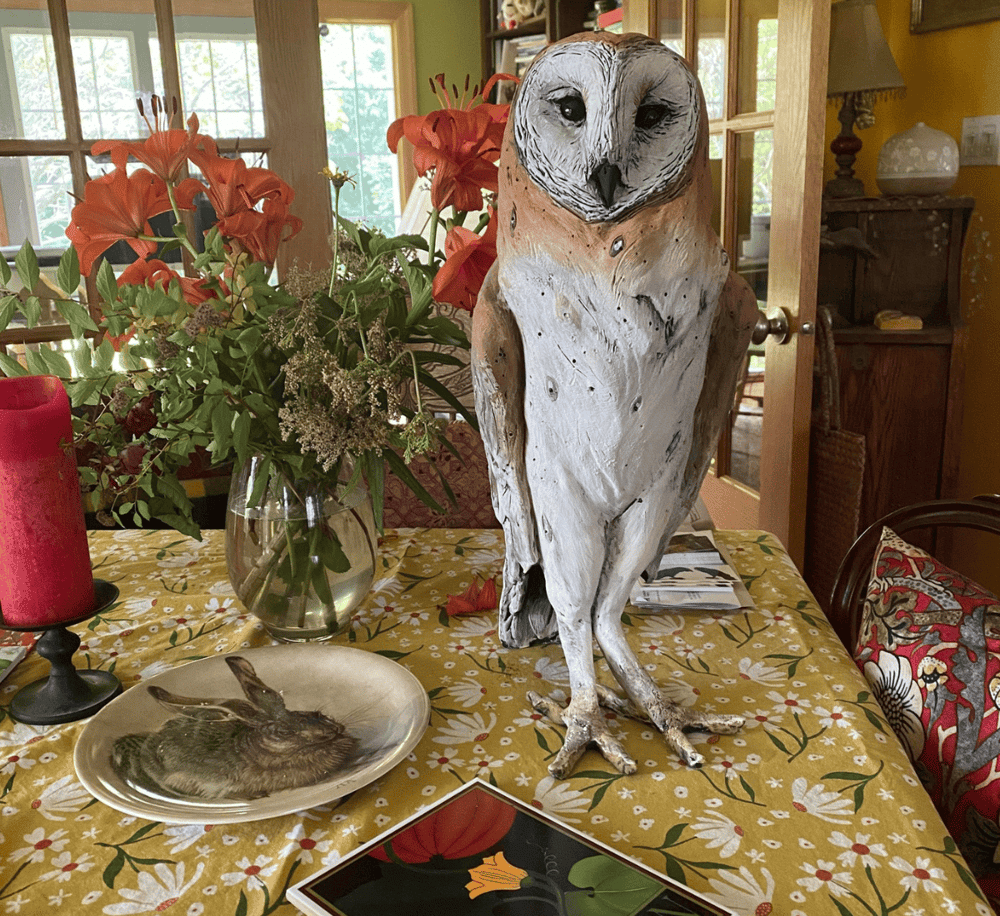 A wood-carved owl sculpture sitting on a floral tablecloth with a flower arrangement and decorative items in a cozy, well-lit room.