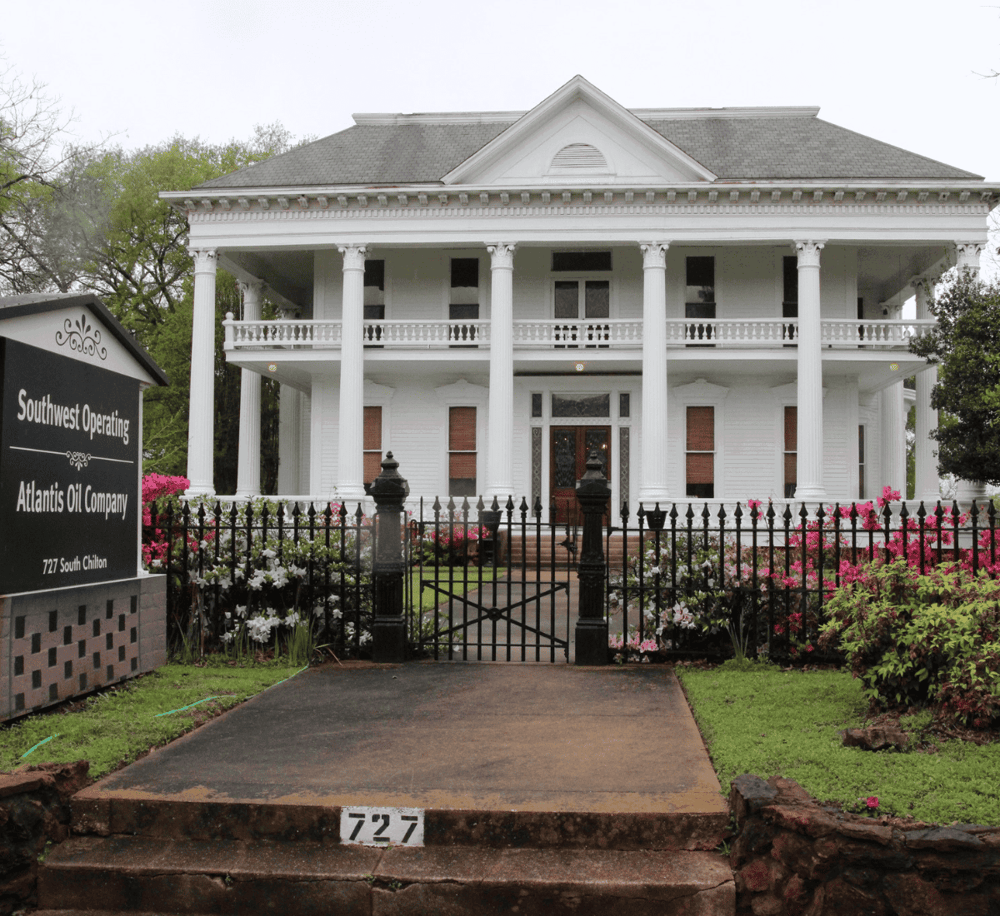 Elegant white historic mansion with large columns, surrounded by colorful flowers and black iron fence.