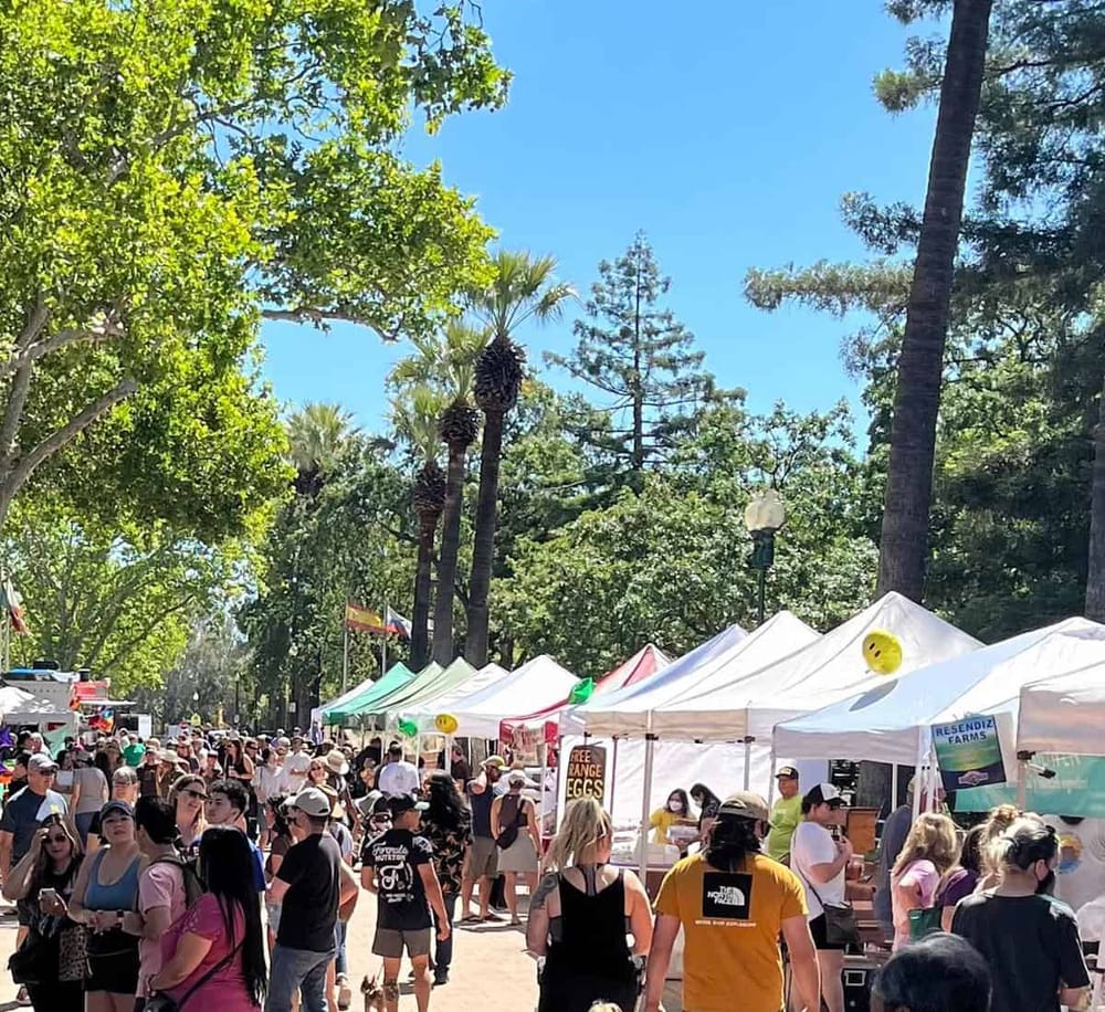 Vibrant outdoor market with tents, shoppers, and lush greenery, perfect for local food and craft shopping.