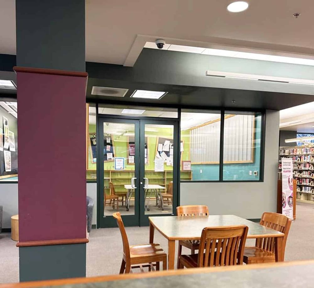 Spacious library reading area with wooden tables and chairs, library entrance with glass doors and bookshelves in the background.