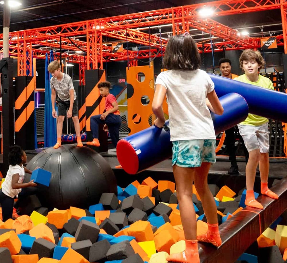 Children playing in an indoor adventure park with obstacle courses and foam pits.