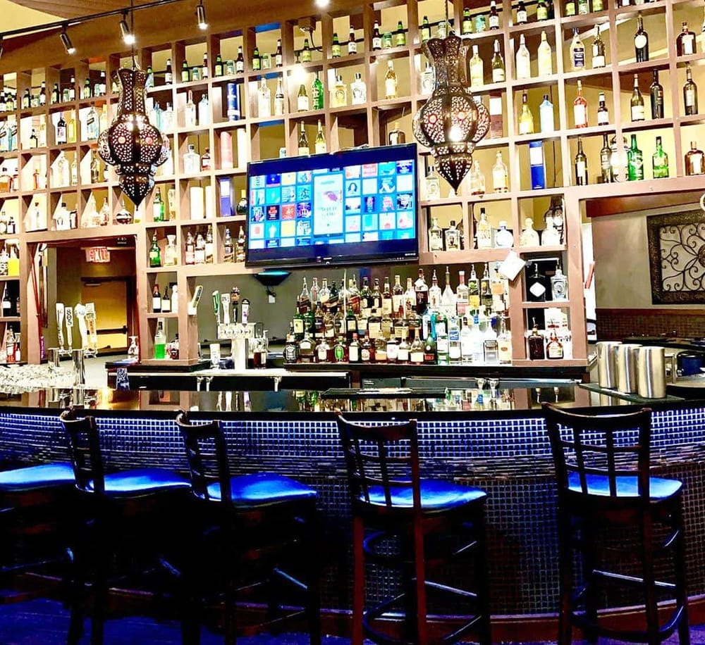 Bright bar with a wide selection of liquors and a digital TV screen displaying streaming content, illuminated by Moroccan-style hanging lamps.