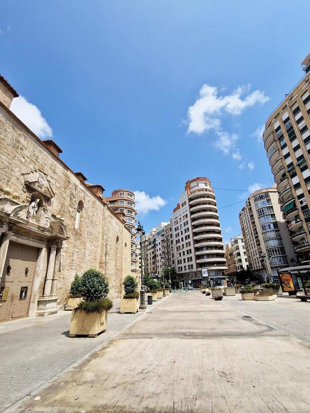Modern city street with historic stone church, high-rise buildings, blue sky, and potted plants, showcasing urban travel destinations.