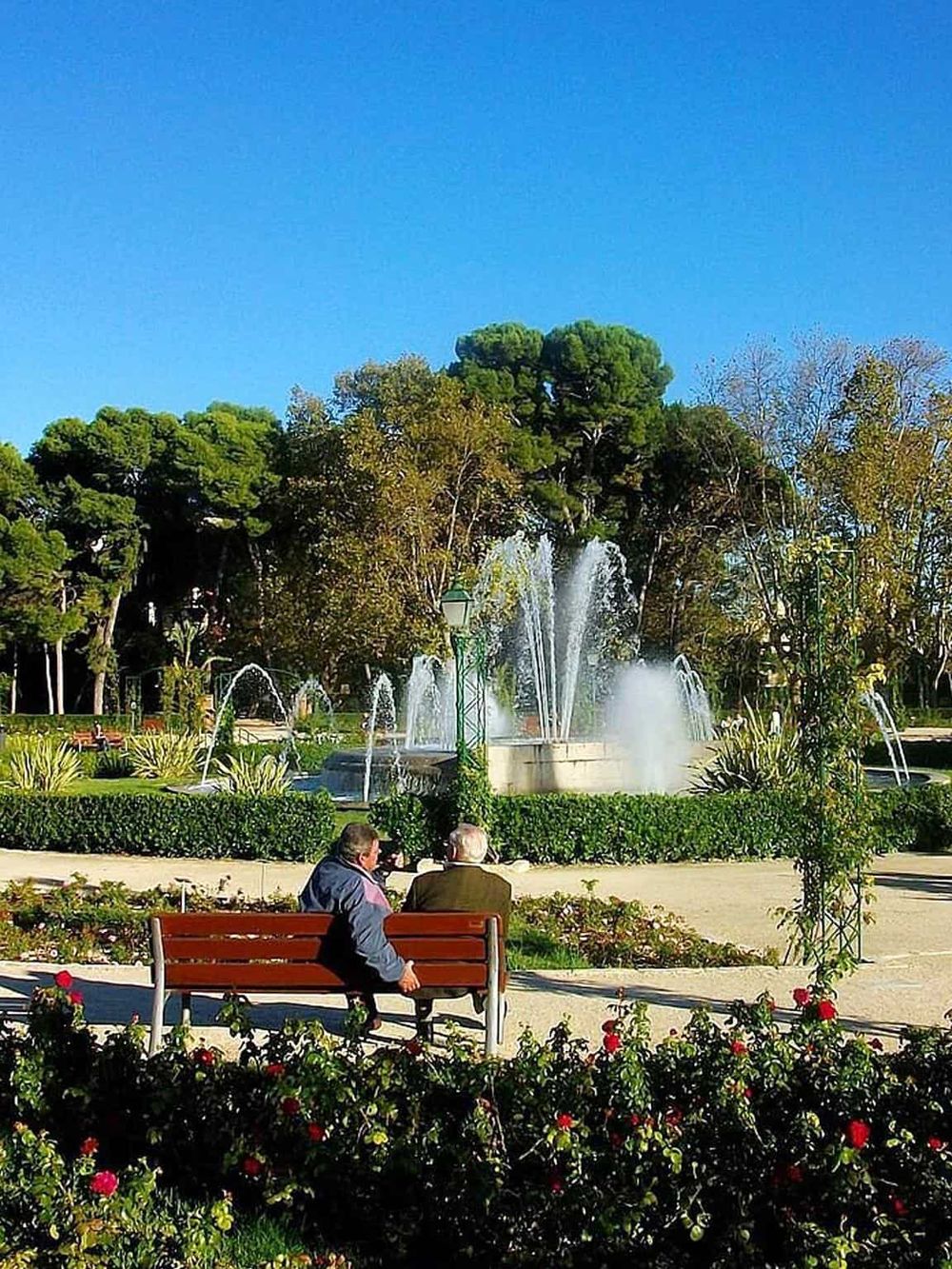 Fountain in a park with two elderly men sitting on a bench under a clear blue sky.