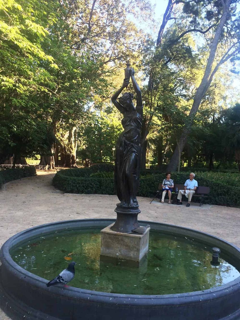 Bronze statue of woman holding a water jug in a park fountain, surrounded by lush trees and visitors enjoying the scenery.