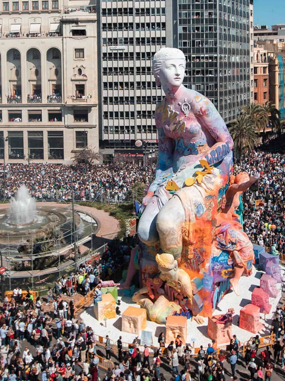 Colorful giant sculpture of a woman surrounded by a dense crowd in an urban city square.