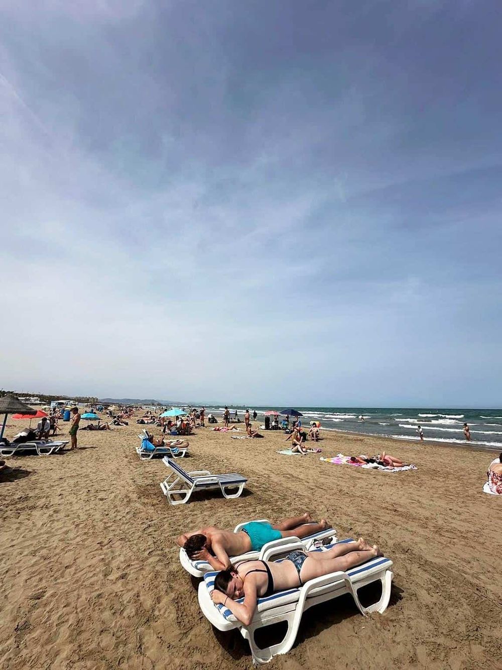 Relaxing beach day with sunbathers on lounge chairs, ocean waves, and clear blue sky, perfect for seaside vacations.