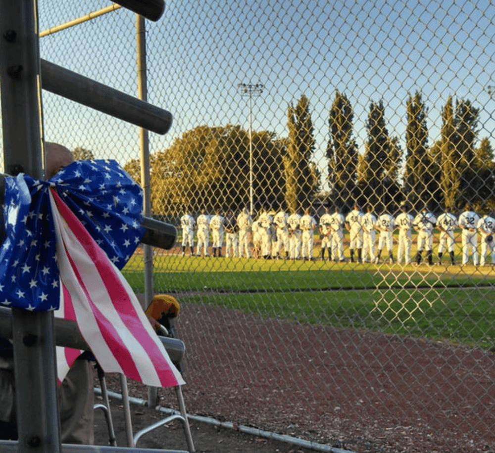 American flag draped over fence at youth baseball game under evening sky.