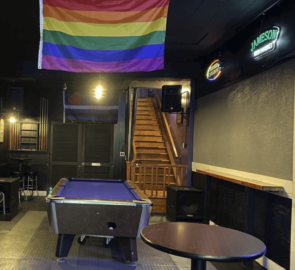 Vibrant rainbow pride flag hanging in cozy indoor bar setting with pool table and staircase.