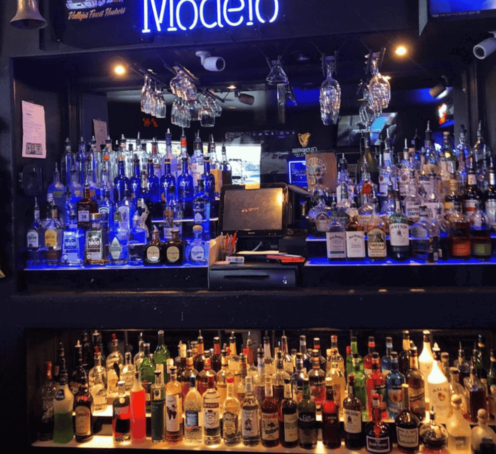 Brightly lit bar with a wide selection of liquor bottles and hanging wine glasses, showcasing a vibrant nightlife scene.