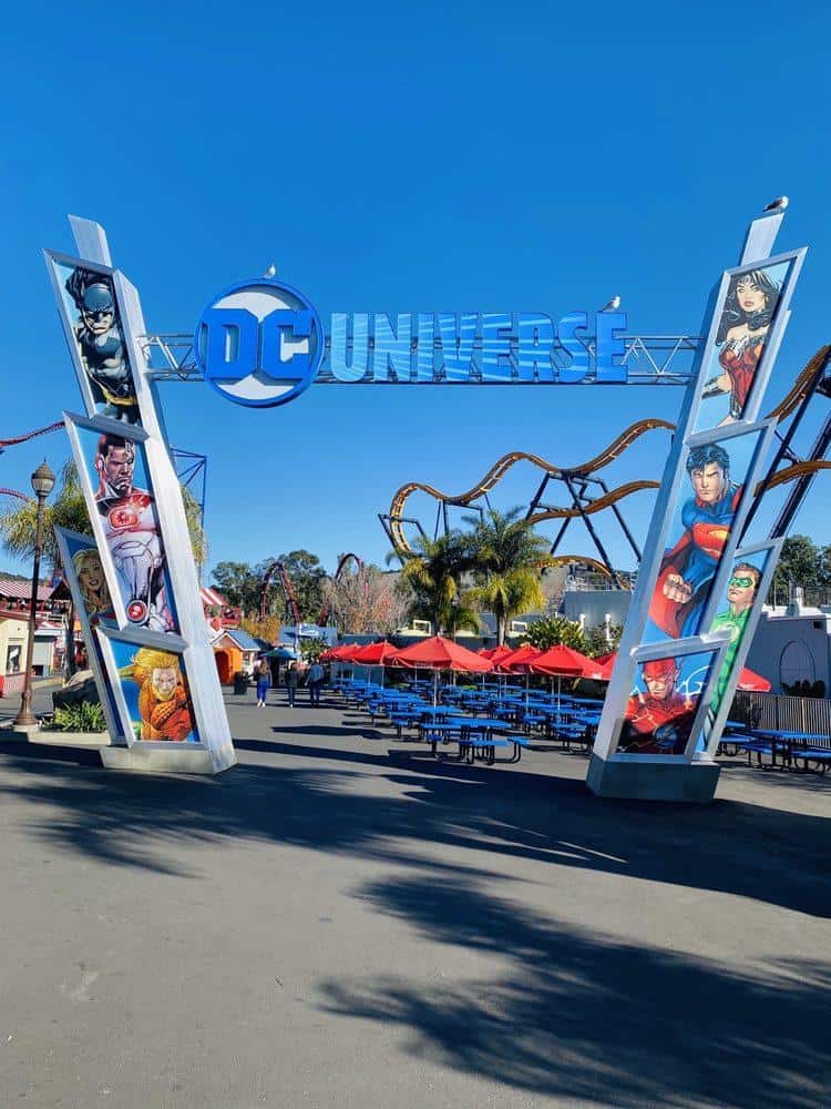 DC Universe theme park entrance with colorful character banners and roller coaster in background.