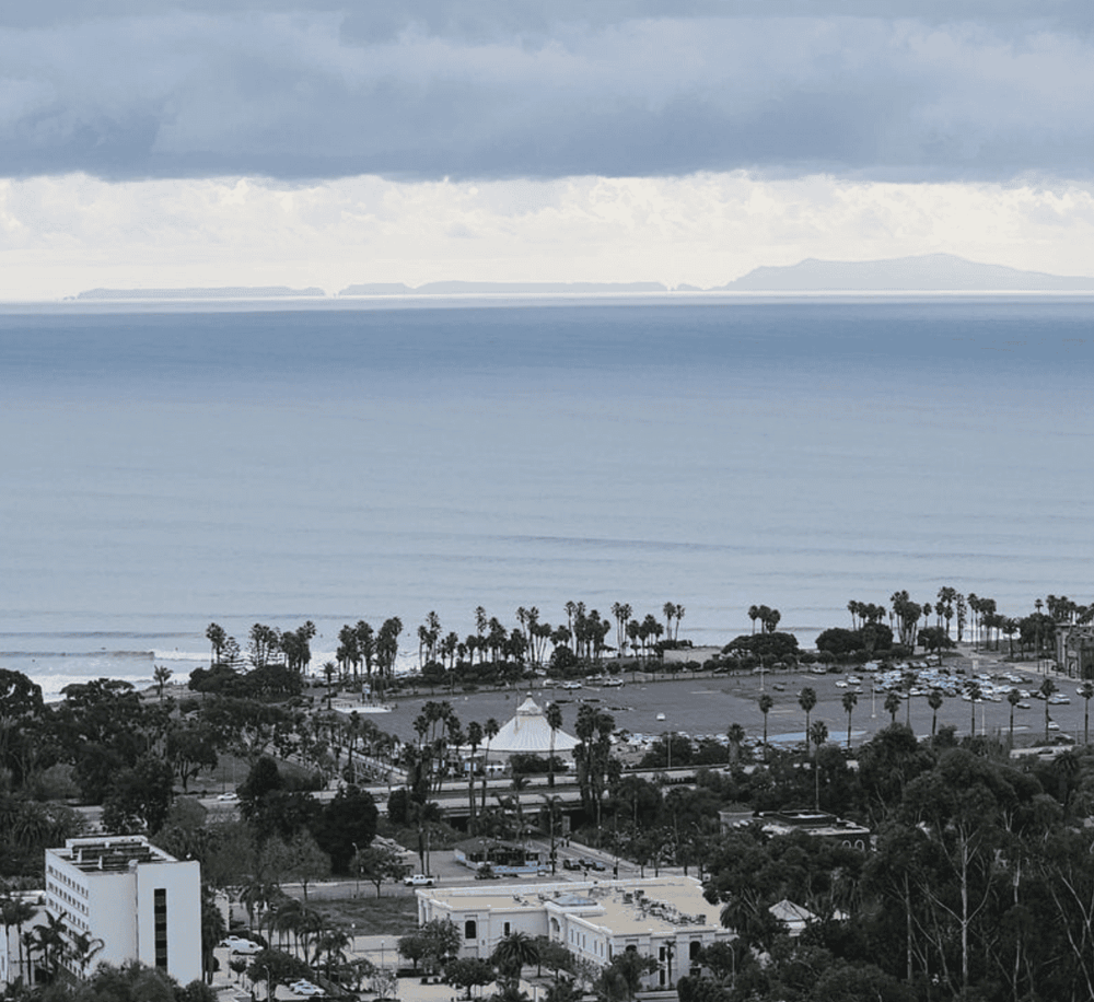 Aerial view of a coastal city with palm trees, parking lots, and ocean scenery, showcasing scenic travel destinations.