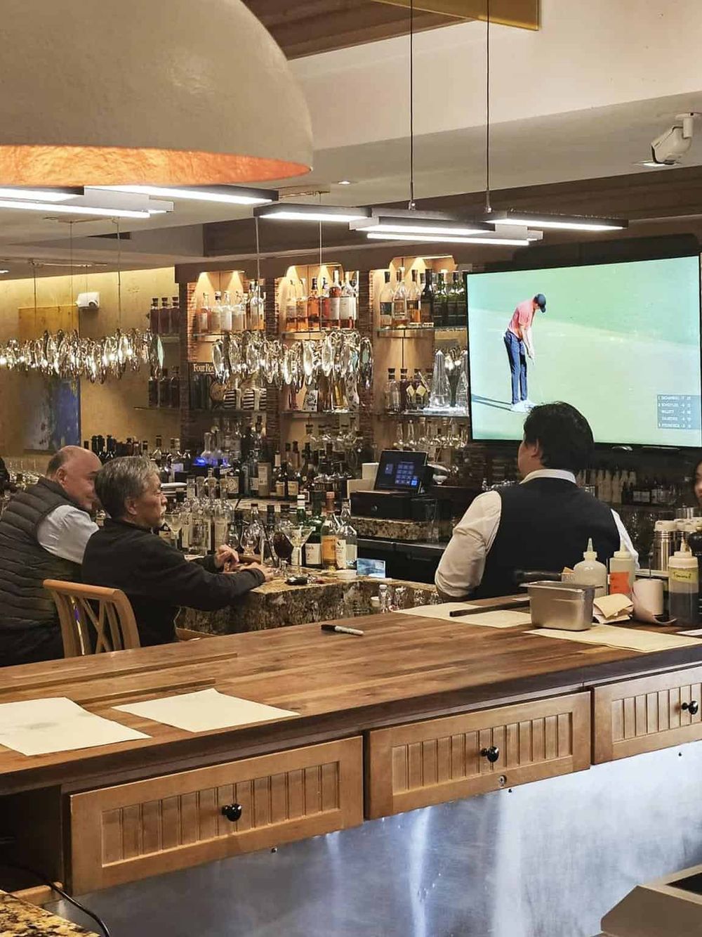 Modern bar interior with people watching golf on large screen TV, alcohol bottles, and stylish decor.