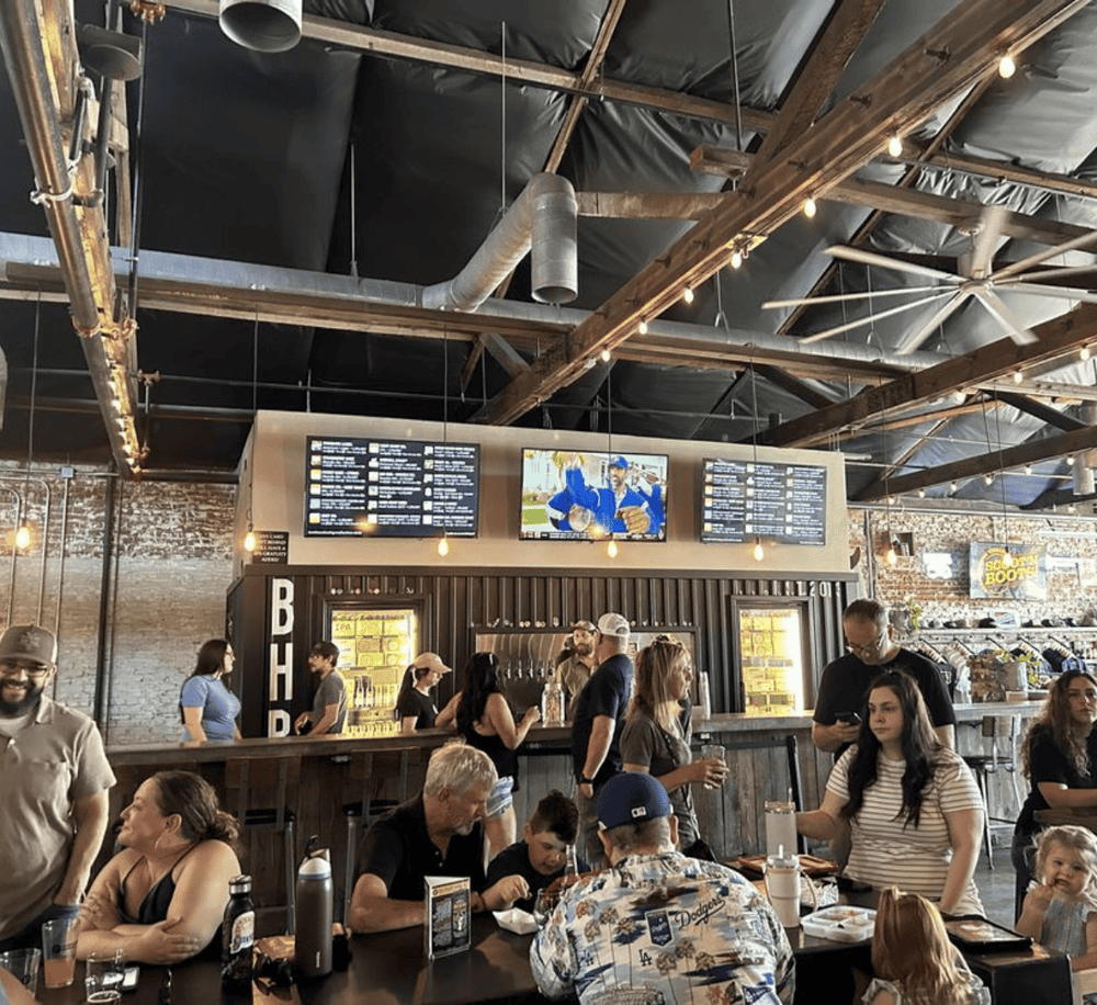 Aerial view of a lively brewery with customers enjoying food and drinks, industrial decor, and large digital menu screens.