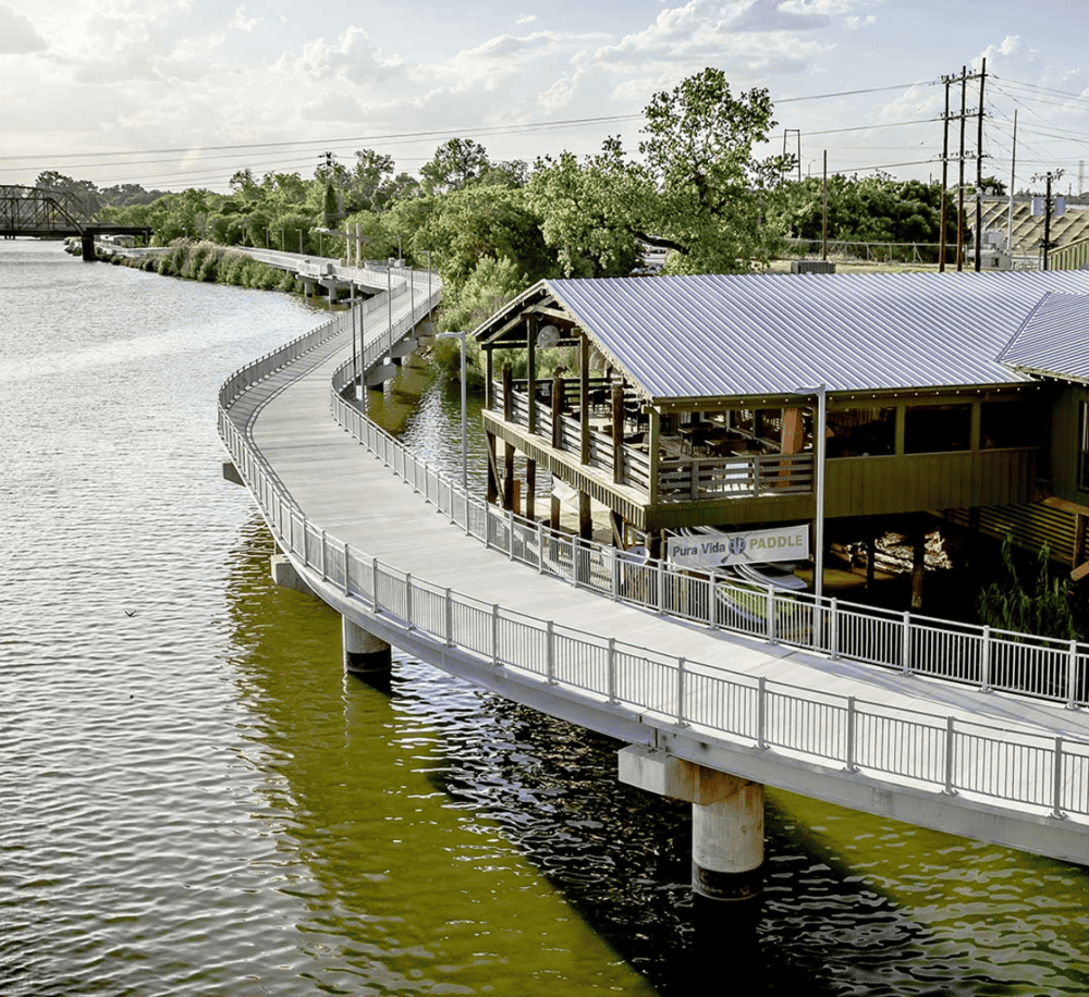 Seaside boardwalk with a restaurant overlooking the water, surrounded by trees and urban infrastructure.