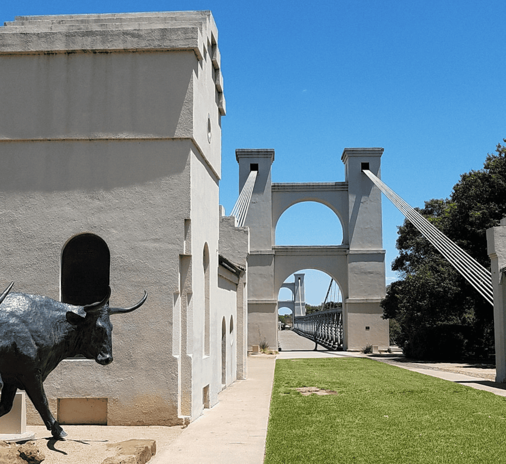 Historic suspension bridge with white arches and a bull statue in front, scenic outdoor location.
