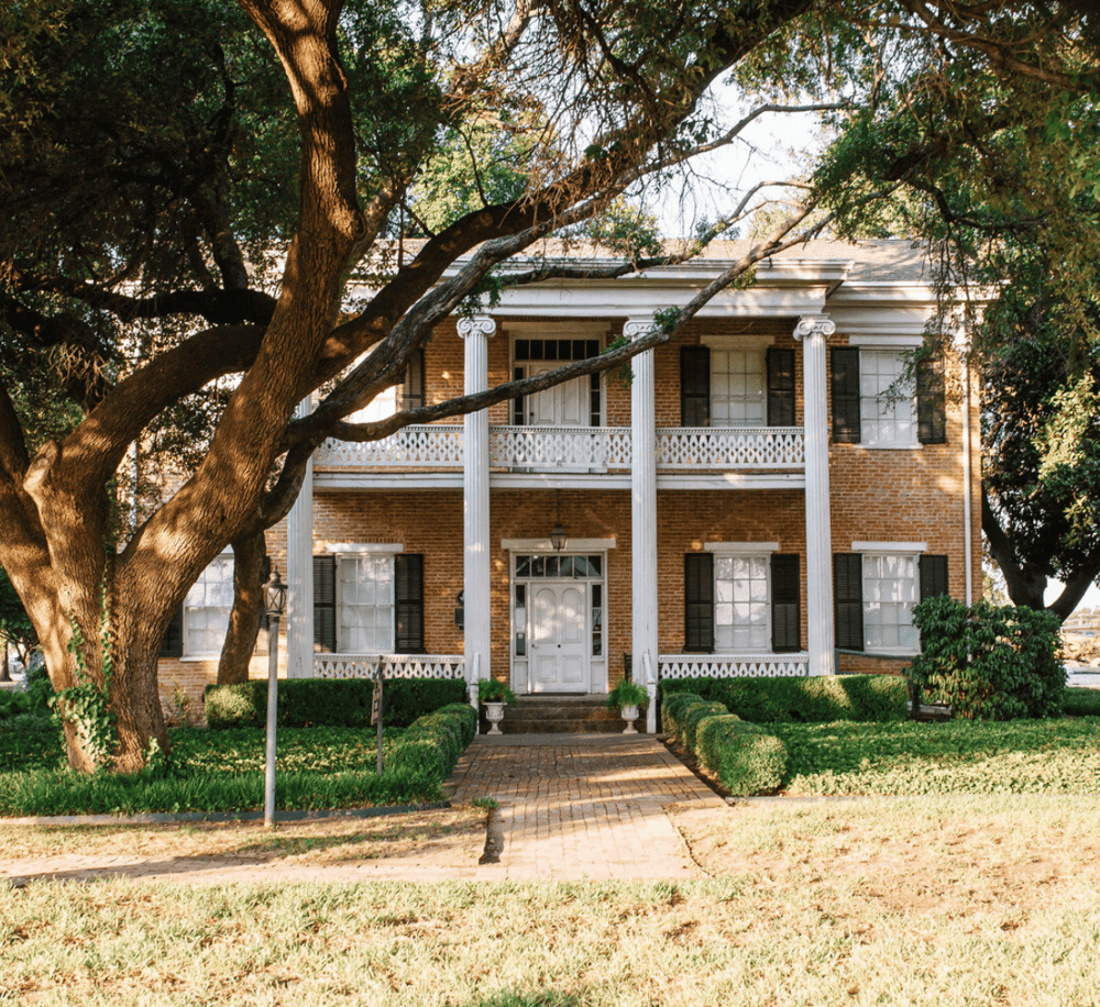 Charming historic brick house with white columns and greenery, ideal for guided home tours.