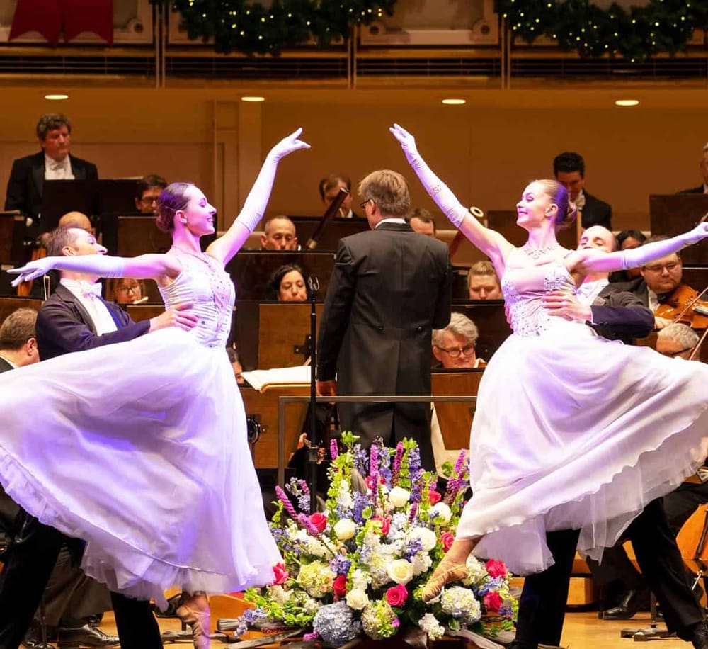 Elegant ballet dancers performing on stage with orchestra in the background.