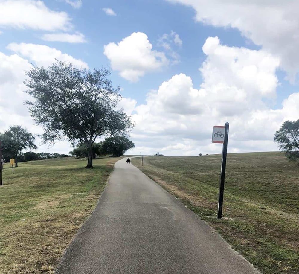 Bicycle path in park with sign, trees, and blue sky with clouds, scenic outdoor recreational area.