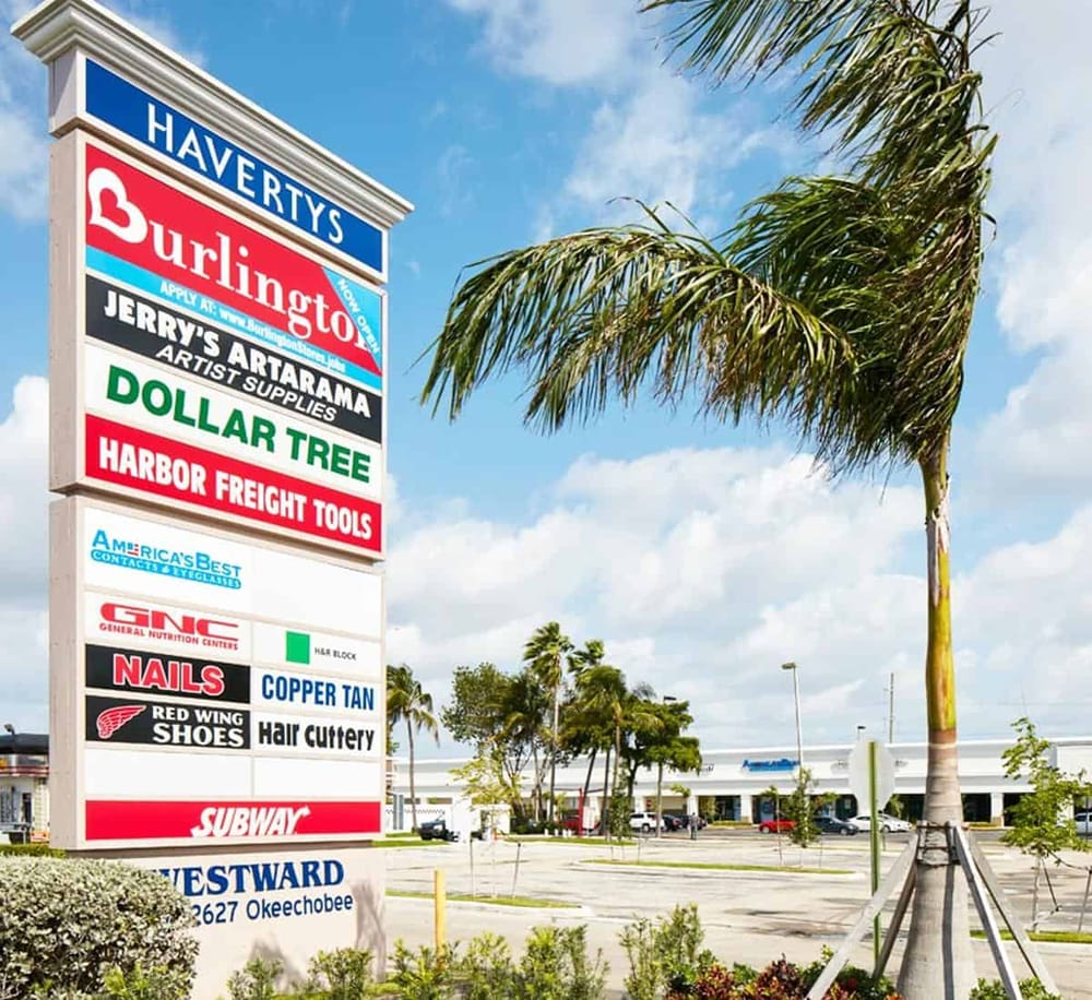 Vast shopping plaza sign with diverse retail stores, palm trees, and blue sky in Okeechobee, Florida.