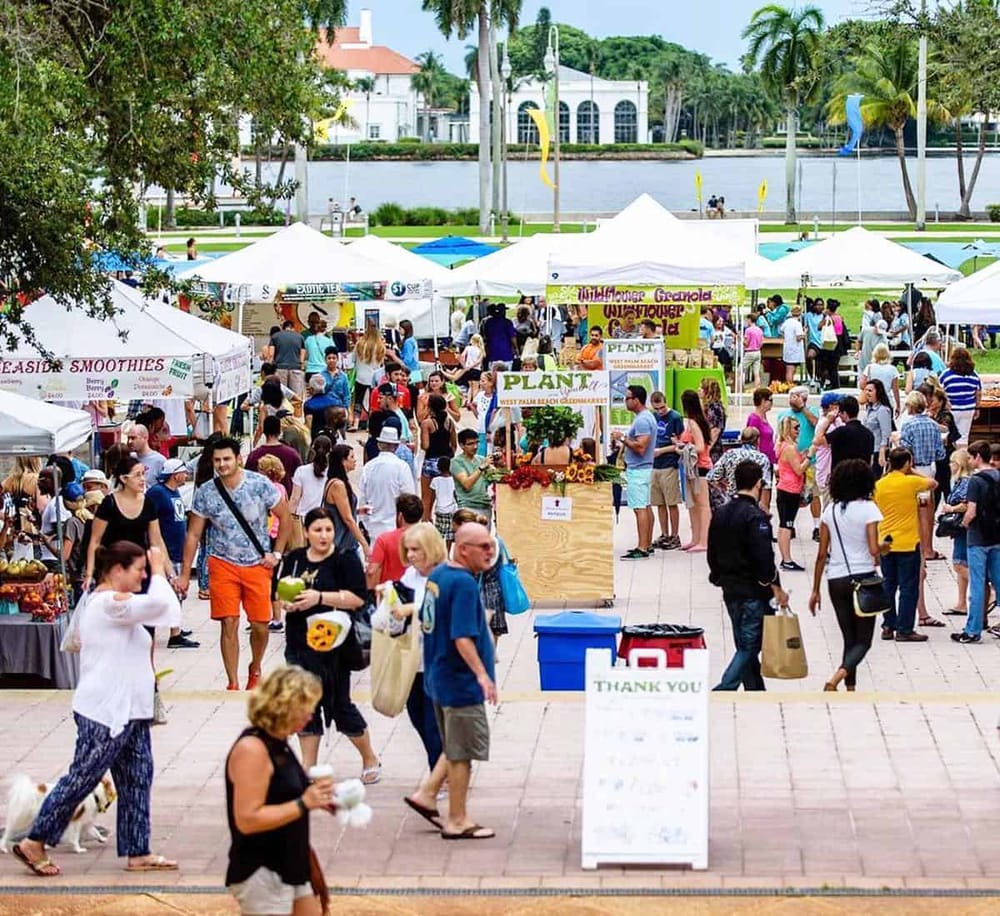 Vibrant outdoor farmers market with food stalls, shoppers, and scenic water backdrop.
