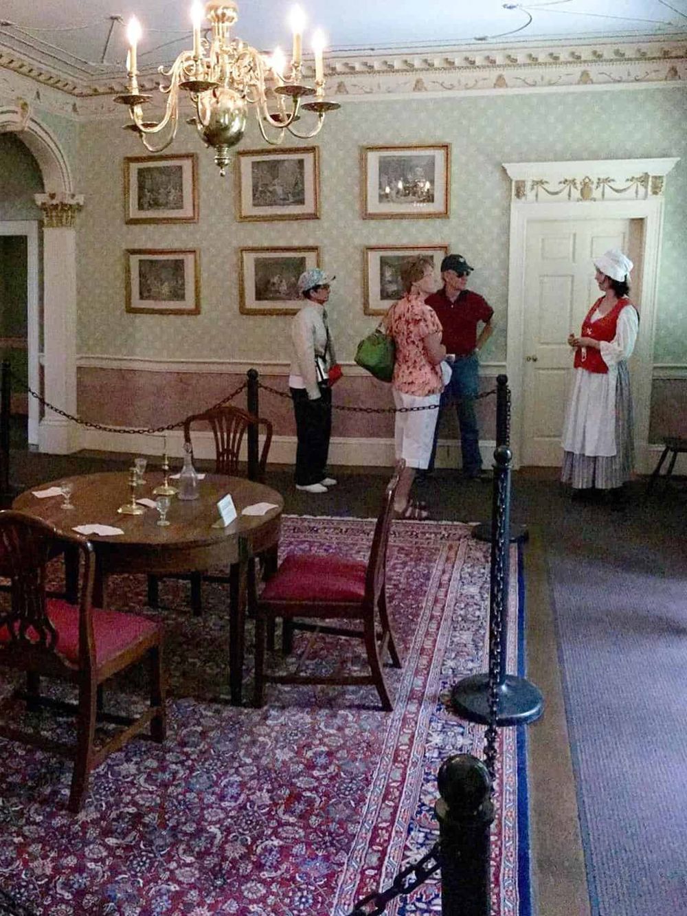 Vintage historical museum room with visitors and guide, elegant chandelier, antique decor, classic artwork, informational tour, cultural heritage site.